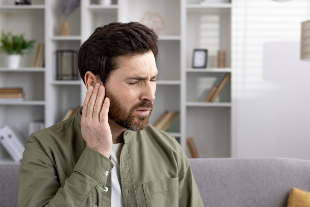 Man experiencing ear pain at home, holding his ear with a pained expression, indicating discomfort and possible inflammation or earache in a peaceful living room setting.